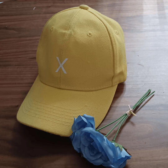 A yellow hat with a white X on the front, sitting next to three blue roses on a wooden table.